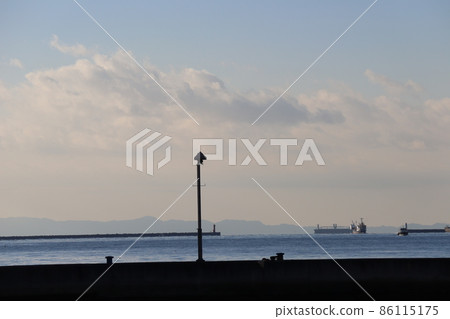 海岸的藍海、碼頭、燈塔、漁船、油輪、朦朧的山、雲和淺藍色的天空景觀，在陰影中看起來是黑色的 86115175