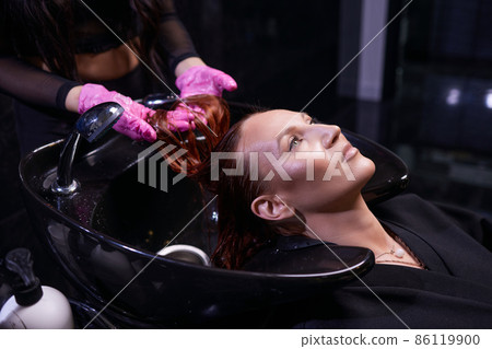 Hairdresser in beauty salon washes his client hair, before procedure of applying natural restoring ingredients and vitamins to hair and haircut. Hairdresser in beauty salon washes his client hair, before procedure of applying natural restoring ingredients and vitamins to hair and haircut. 86119900