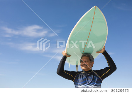 Low angle view of biracial senior man carrying surfboard over head at sunny beach against blue sky Low angle view of biracial senior man carrying surfboard over head at sunny beach against blue sky 86120475