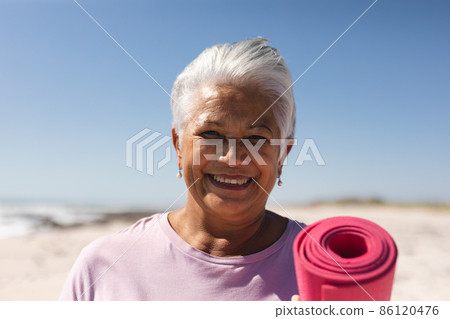 Portrait of smiling retired biracial senior woman holding exercise mat with short hair at beach 86120476