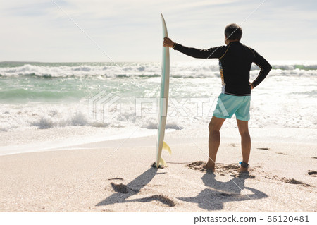 Full length rear view of biracial senior man holding surfboard looking at waves in sea on sunny day 86120481