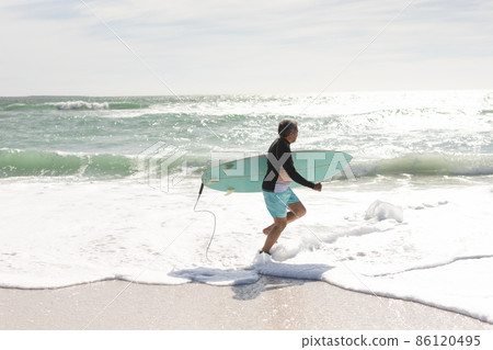 Side view of biracial senior man carrying surfboard running on shore at beach during sunny day 86120495