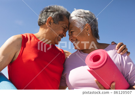 Happy biracial senior man and woman touching foreheads holding yoga mats at beach on sunny day 86120507