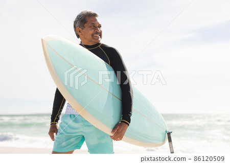 Smiling biracial senior man carrying surfboard looking away at beach against sky during sunny day 86120509