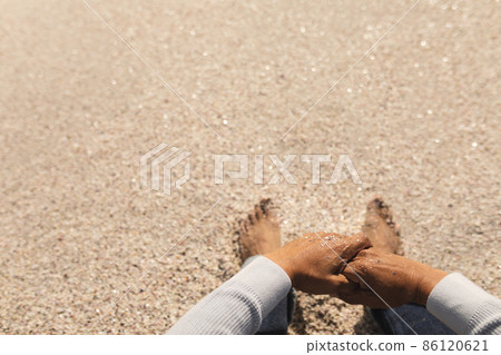 Low section of biracial senior man sitting with hands clasped on sand at beach during sunny day 86120621