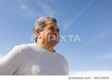 Low angle view of smiling biracial senior man looking away at beach against blue sky on sunny day Low angle view of smiling biracial senior man looking away at beach against blue sky on sunny day 86120644