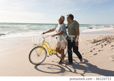 Biracial senior man walking and talking with woman wheeling bicycle at sunny beach against sky 86120715