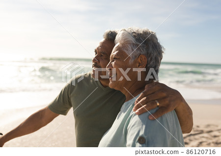 Biracial senior man with arm around woman standing by bicycle looking away at beach on sunny day 86120716