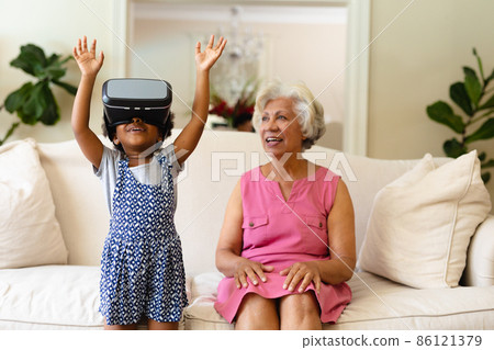 African american grandmother looking at her granddaughter wearing vr headset at home African american grandmother looking at her granddaughter wearing vr headset at home 86121379