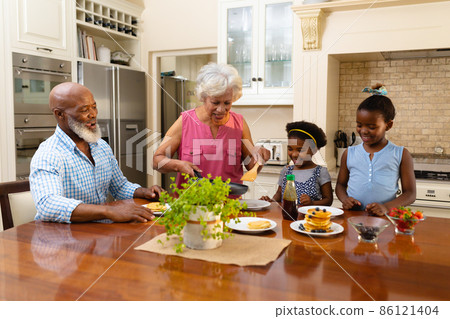 African american grandmother serving breakfast for her two granddaughters in kitchen at home African american grandmother serving breakfast for her two granddaughters in kitchen at home 86121404