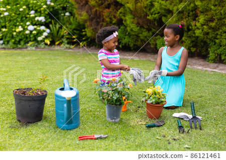 Playful smiling african american sisters kneeling while wearing gloves by plants in garden 86121461