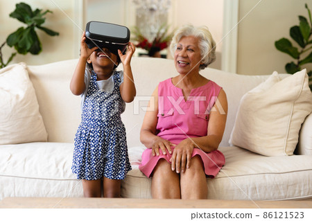 African american girl wearing vr headset and grandmother smiling sitting on the couch 86121523