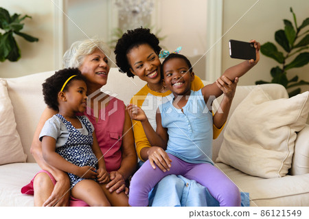 African american grandmother, mother and two granddaughters taking a selfie sitting on couch 86121549