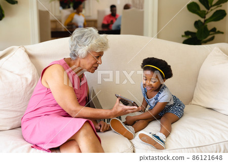 African american grandmother showing digital tablet to her granddaughter sitting on couch at home African american grandmother showing digital tablet to her granddaughter sitting on couch at home 86121648