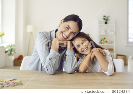 Happy teacher and student girl sitting at working desk in classroom. Cheerful young mother and child sitting at table and smiling. Little kid and her private language tutor looking at camera together 86124284