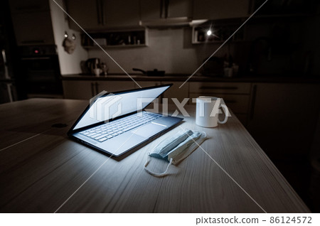 Remote work overtime during the quarantine period in the home office. Working atmosphere in the kitchen on a laptop in the dark late at night. Coffee mug and medical mask on the table. 86124572