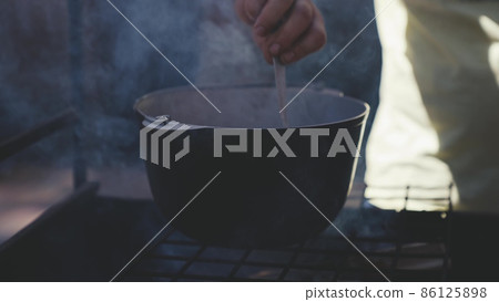 a man cooks food in a pot on fire, delicious cooking in a saucepan in a tourist camp, a recipe on coals in smoke, a trip to the forest to nature, an actively spent weekend, cook stirs food with spoon a man cooks food in a pot on fire, delicious cooking in a saucepan in a tourist camp, a recipe on coals in smoke, a trip to the forest to nature, an actively spent weekend, cook stirs food with spoon 86125898