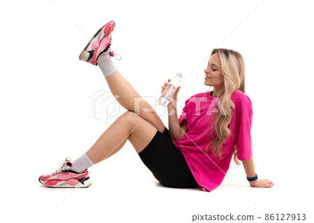 young woman in sportswear sits on a white isolated background and holds a bottle of water in her 86127913
