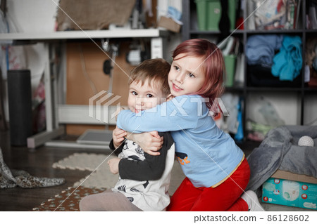Little caucasian girl hugging with her brother on the floor in their room Little caucasian girl hugging with her brother on the floor in their room 86128602