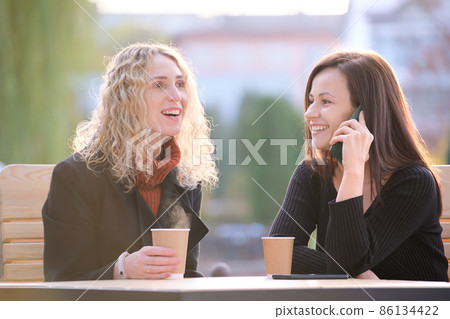Two happy women friends having fun time together at street cafe laughing and talking on cellphone Two happy women friends having fun time together at street cafe laughing and talking on cellphone 86134422
