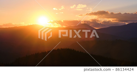 Beautiful evening panoramic landscape with bright setting sun over distant mountain peaks at sunset Beautiful evening panoramic landscape with bright setting sun over distant mountain peaks at sunset 86134424
