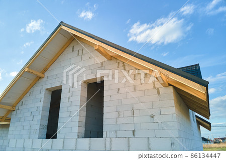 Aerial view of unfinished house with aerated lightweight concrete walls and wooden roof frame covered with metallic tiles under construction. Aerial view of unfinished house with aerated lightweight concrete walls and wooden roof frame covered with metallic tiles under construction. 86134447