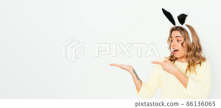 Cheerful surprised young woman wearing bunny ears pointing on empty space 86136065