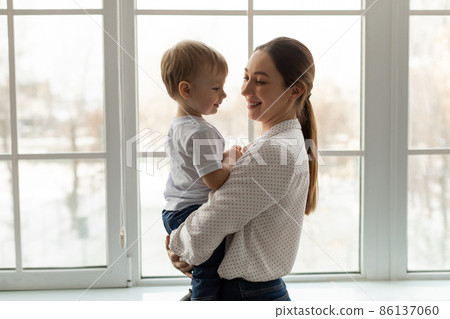 Mom's love. Young mom holding toddler son on hands, cuddling with kid boy, standing near window indoors, free space Mom's love. Young mom holding toddler son on hands, cuddling with kid boy, standing near window indoors, free space 86137060
