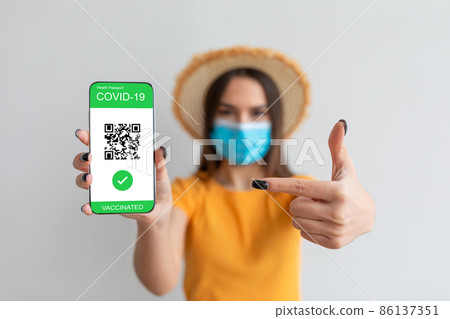 Woman pointing at smartphone with digital covid-19 vaccination certificate on screen, lady wearing medical mask 86137351