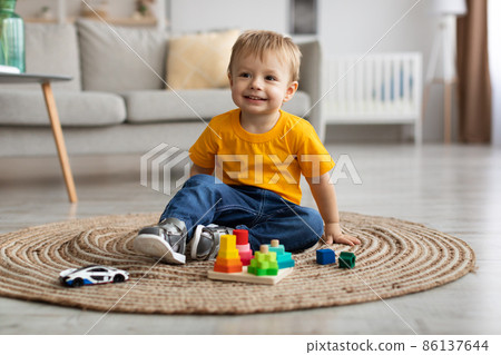 Cute toddler boy playing with car and educational wooden toy at home, sitting on floor carpet in living room and smiling Cute toddler boy playing with car and educational wooden toy at home, sitting on floor carpet in living room and smiling 86137644