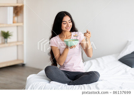 Full length portrait of beautiful millennial Indian lady having cereal with milk while sitting on bed at home 86137699
