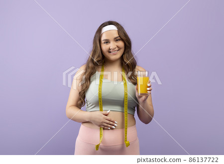 Happy pretty caucasian plus size young woman in sportswear with measuring tape show glass of fresh orange juice Happy pretty caucasian plus size young woman in sportswear with measuring tape show glass of fresh orange juice 86137722