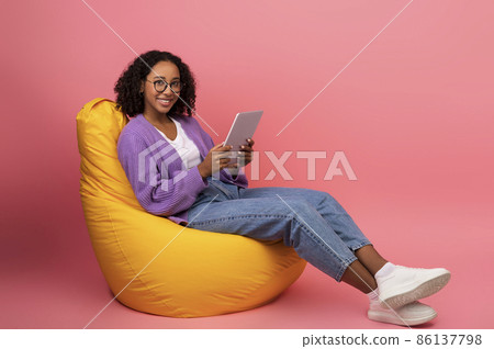 Full length of joyful young black woman with digital tablet sitting on bean bag chair, working online on pink background Full length of joyful young black woman with digital tablet sitting on bean bag chair, working online on pink background 86137798