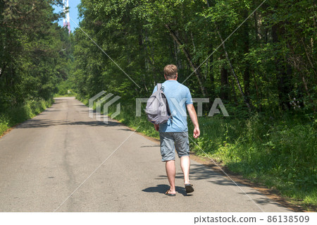 An adult male traveler with a backpack is walking alone on an empty road in the middle of the forest An adult male traveler with a backpack is walking alone on an empty road in the middle of the forest 86138509