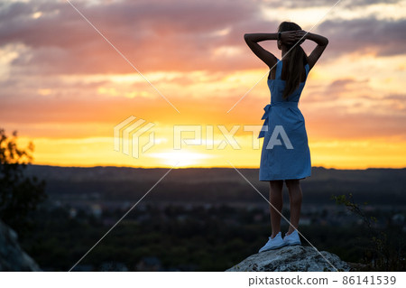 A young woman in summer dress standing outdoors enjoying view of bright yellow sunset. A young woman in summer dress standing outdoors enjoying view of bright yellow sunset. 86141539