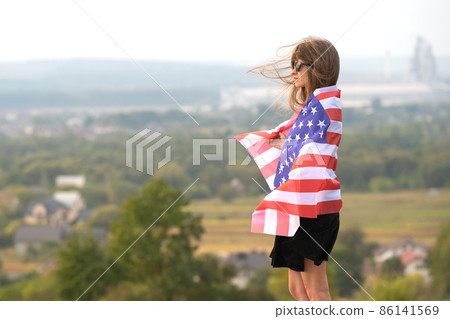 Young pretty american woman with long hair holding waving on wind USA flag on her sholders standing outdoors enjoying warm summer day. Young pretty american woman with long hair holding waving on wind USA flag on her sholders standing outdoors enjoying warm summer day. 86141569