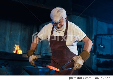 Focused craftsman in apron and gloves forging steel  86142836