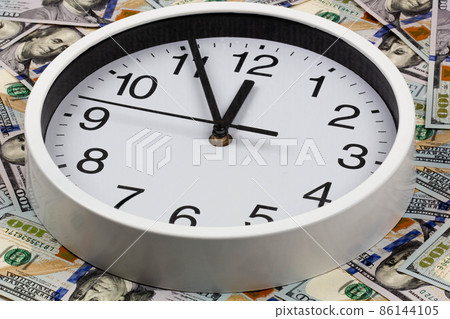 Time is money. A large white watch with black numbers is surrounded by hundred dollar bills. Business and finance concept. The clock shows the time at five minutes to twelve on US dollars background Time is money. A large white watch with black numbers is surrounded by hundred dollar bills. Business and finance concept. The clock shows the time at five minutes to twelve on US dollars background 86144105