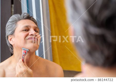 An adult gray-haired man shaves with a razor in front of a mirror 86144722