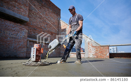 Side view of young worker in protective gloves leveling concrete slab using special machine. Concept of process construction new modern house with special equipments outdoors. 86145180