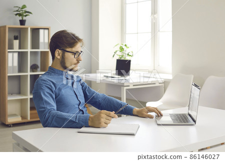 Young Caucasian man in glasses sit at desk work online on laptop in office make notes. Millennial male study on computer, take course or training on internet. Distant education, technology concept. 86146027