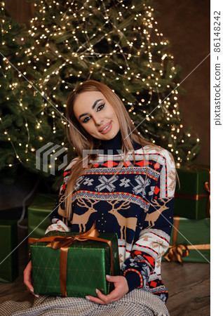 A girl in a sweater with a deer sits on the floor and holds a green gift box. Christmas trees in the background A girl in a sweater with a deer sits on the floor and holds a green gift box. Christmas trees in the background 86148242