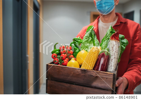 Delivery man in protective mask holding paper bag with food in the entrance. The courier gives the box with fresh vegetables and fruits to the customer 86148901