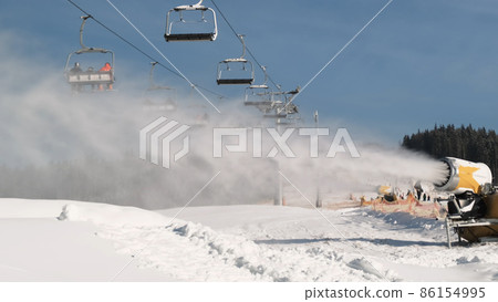 Snow cannon making snow by daytime. Ski lift with skiers is moving in background, skiers and snowboarders drive down the snow covered slope. Snowmaker produces a lot artificial snow Snow cannon making snow by daytime. Ski lift with skiers is moving in background, skiers and snowboarders drive down the snow covered slope. Snowmaker produces a lot artificial snow 86154995
