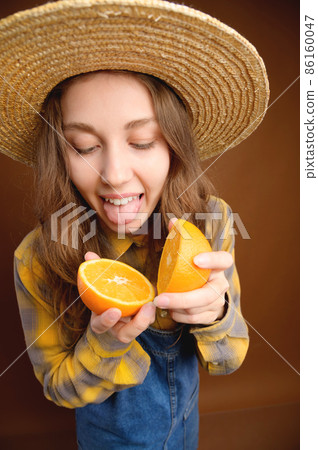 Studio portrait of an attractive caucasian young woman in a hat and denim overalls holds a cut orange fruit in her hands and shows her tongue trying to taste it Studio portrait of an attractive caucasian young woman in a hat and denim overalls holds a cut orange fruit in her hands and shows her tongue trying to taste it 86160047
