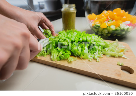 Female hands cut lettuce leaves with a knife against the background of a bowl with chopped vegetables. Vegetarian food preparation 86160050