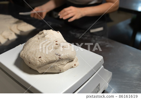 Close-up on a production scale is a portion of bread dough. Hands of a woman baker in defocus in the background. Bread production in a bakery 86161619