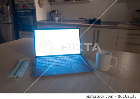 The kitchen is flat. The working atmosphere in the apartment. Laptop in the dark late at night. Work overtime in the home office. Mug and medical mask on the table. 86162133