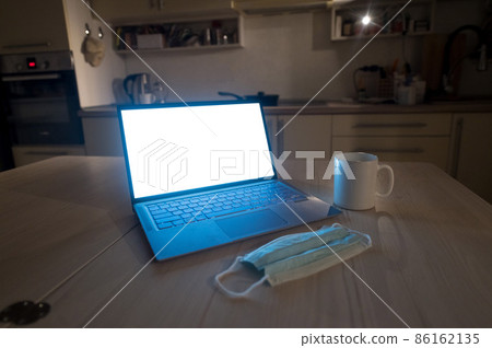 The kitchen is flat. The working atmosphere in the apartment. Laptop in the dark late at night. Work overtime in the home office. Mug and medical mask on the table. 86162135