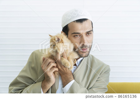 Handsome muslim young man holding lovely brown cut furry kitten sitting together on yellow sofa Handsome muslim young man holding lovely brown cut furry kitten sitting together on yellow sofa 86165136